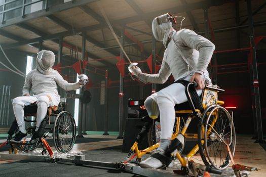 Two athletes in wheelchairs practicing fencing in an indoor gym.