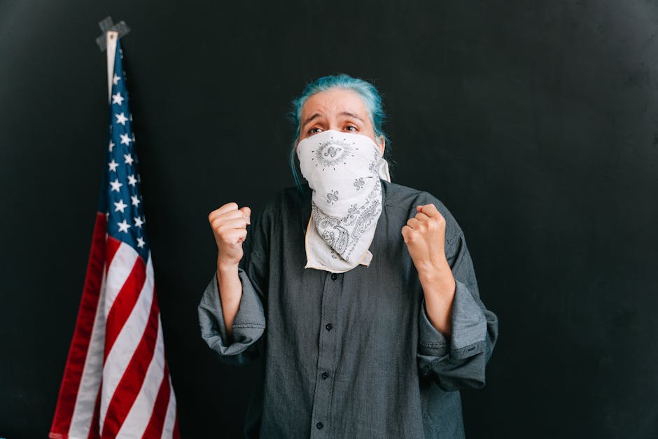 A woman passionately protesting with clenched fists, wearing a bandana mask, next to an American flag.