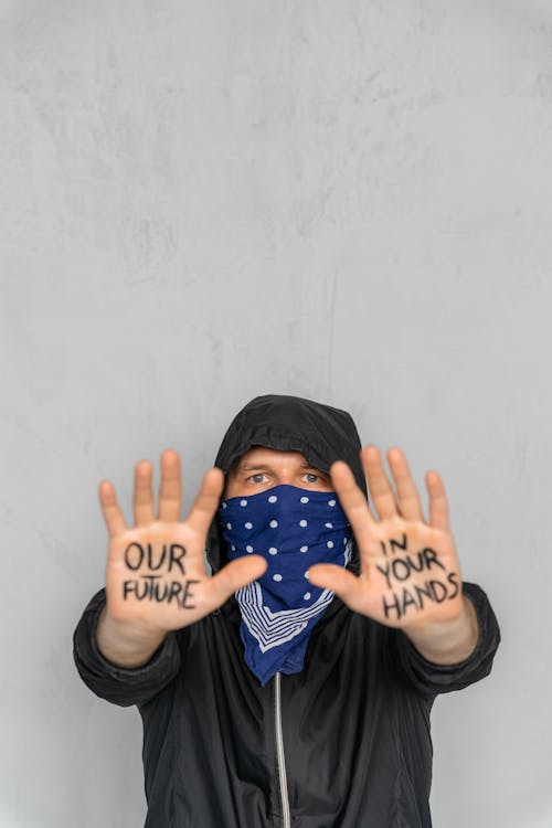 Man Wearing a Hood and Bandana on His Face Showing His Hands with a Message Written on Them 
