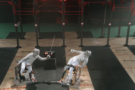 Two fencers in wheelchairs engage in a competitive match indoors, showcasing adaptive sports.