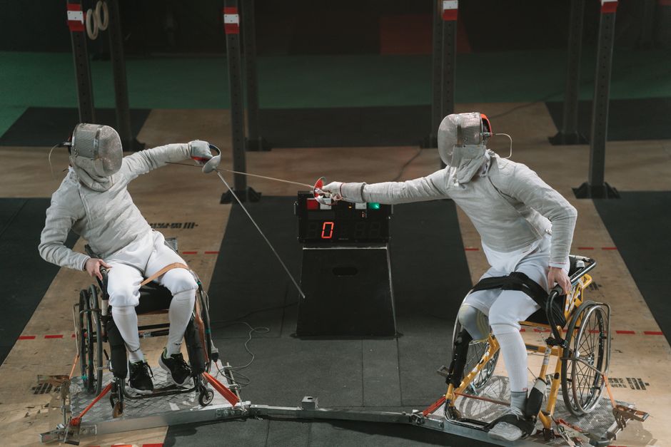 Two male wheelchair fencers in protective gear compete indoors, showcasing dynamic sportsmanship.
