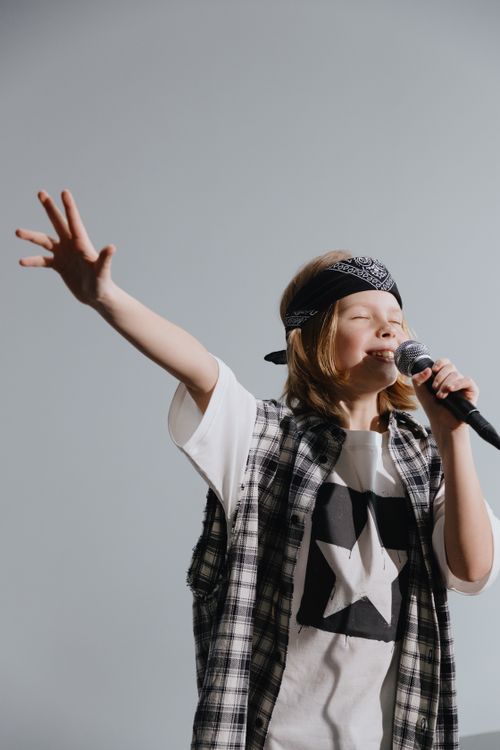 Free Joyful child singing with a microphone, wearing a bandana and plaid shirt, against a neutral background. Stock Photo
