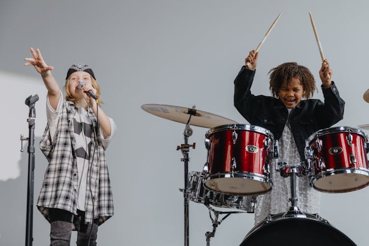 Photograph Of A Kid Playing The Drums Beside A Child Singing