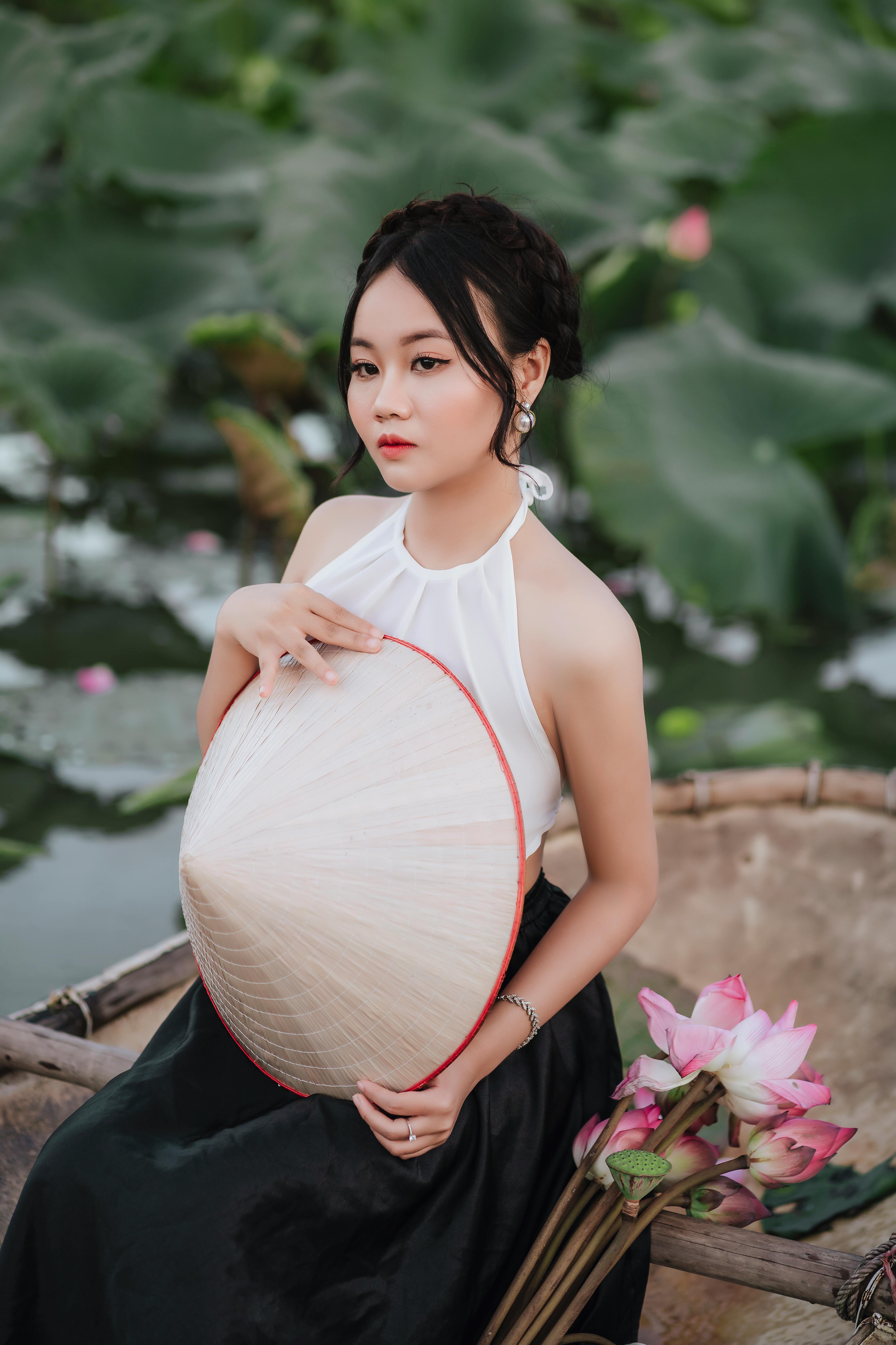 An elegant woman holds a conical hat by a serene lotus pond, exuding grace and beauty.