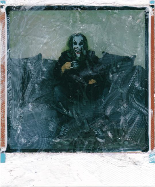 A creepy figure with joker makeup sits on a couch, captured in a vintage Polaroid style.