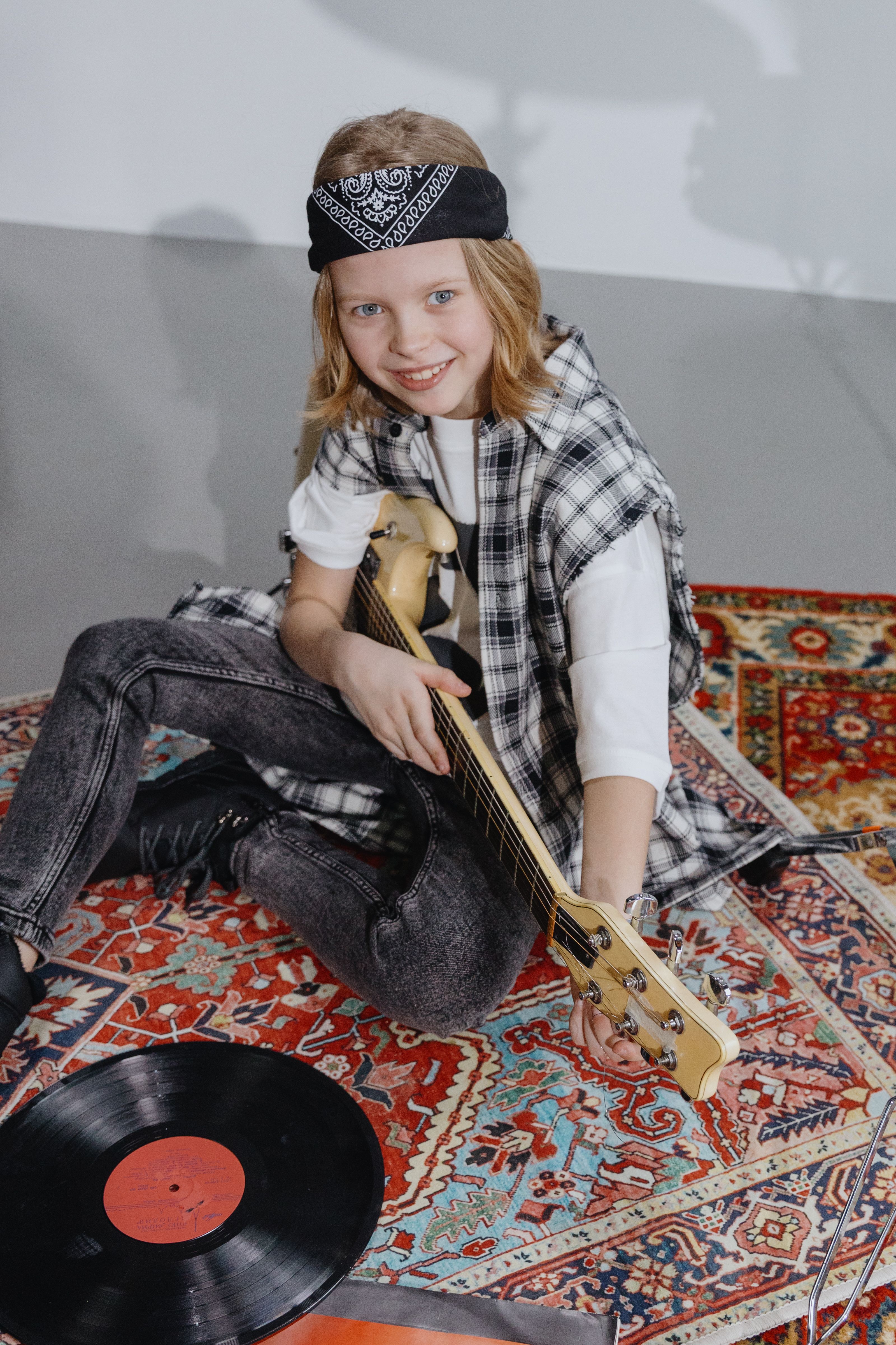 Child with electric guitar smiling on patterned rug next to vinyl record.