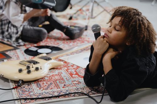 Free Adorable child with curly hair holds microphone on a colorful rug next to a guitar and vinyl record. Stock Photo