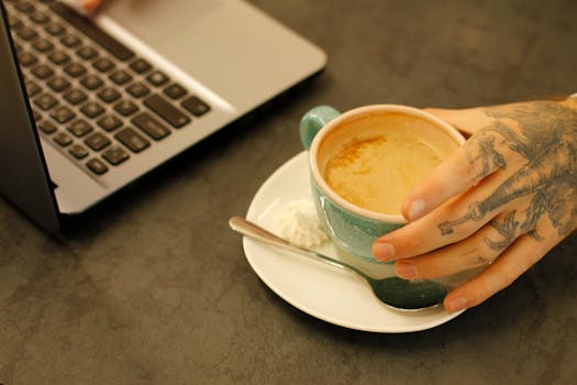 A tattooed hand holds a coffee cup beside a laptop in a cozy cafe setting.