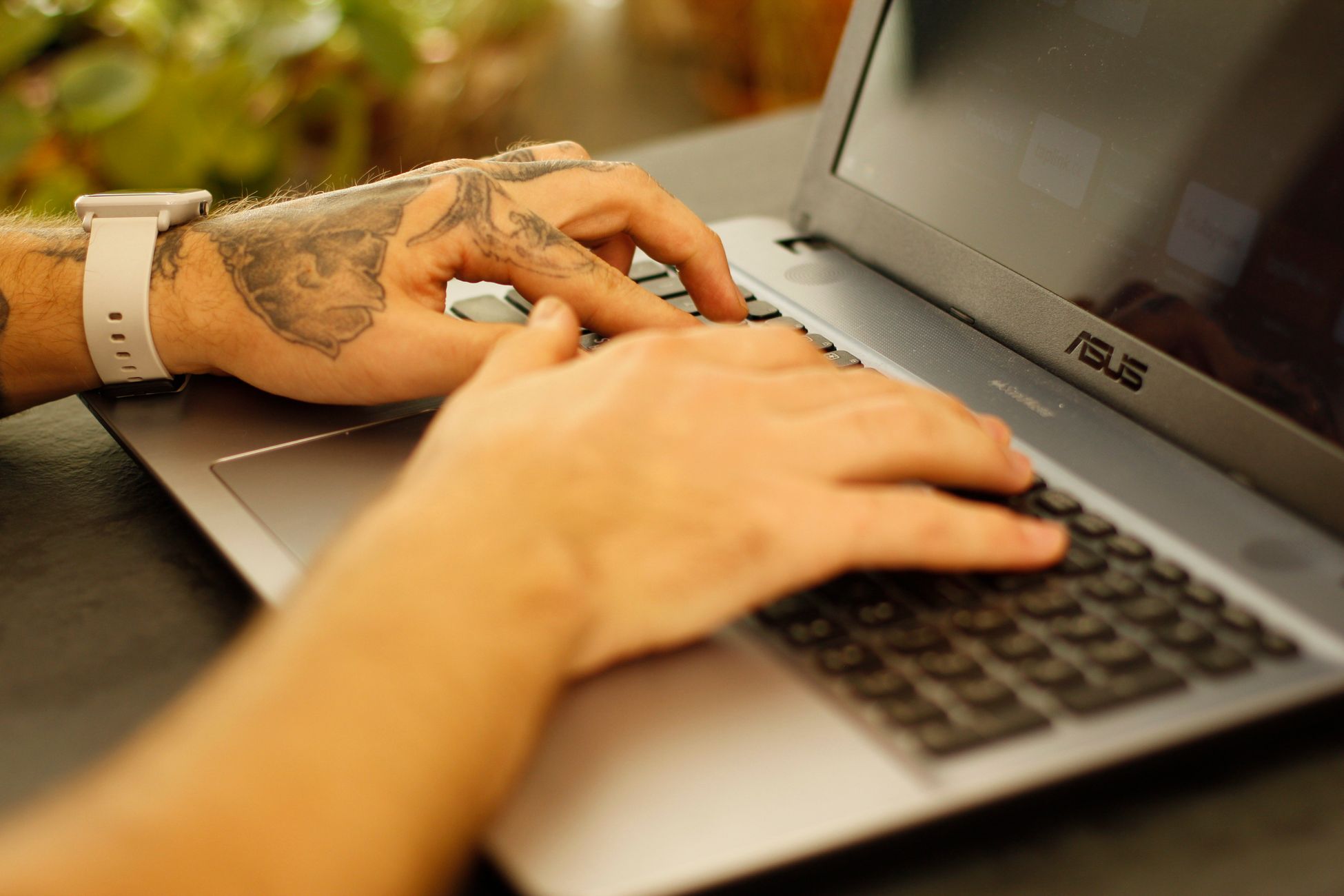 Tattooed hands typing on a laptop keyboard, focused and working.
