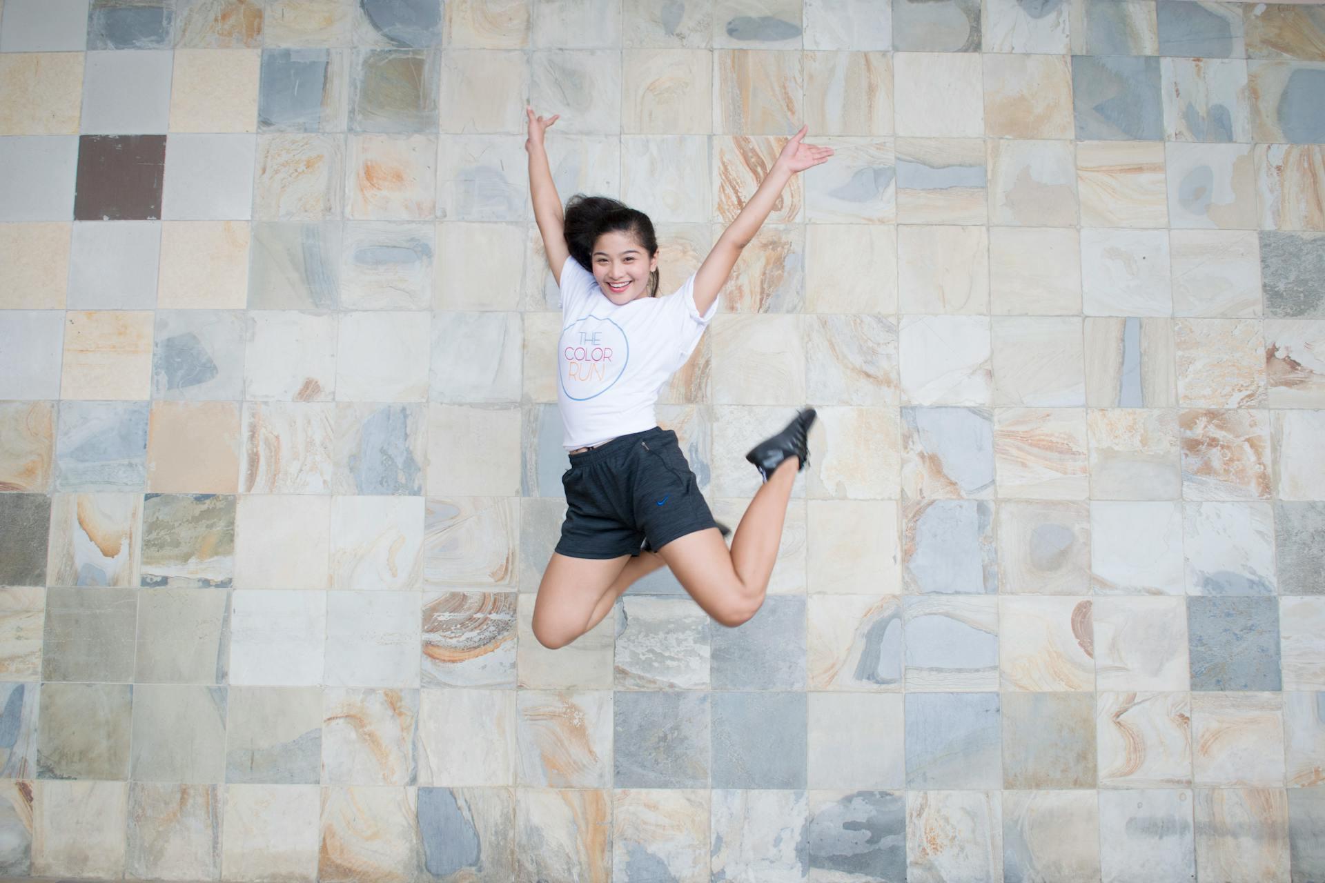 Une jeune femme saute joyeusement contre un mur en carreaux, exprimant son bonheur et son énergie.