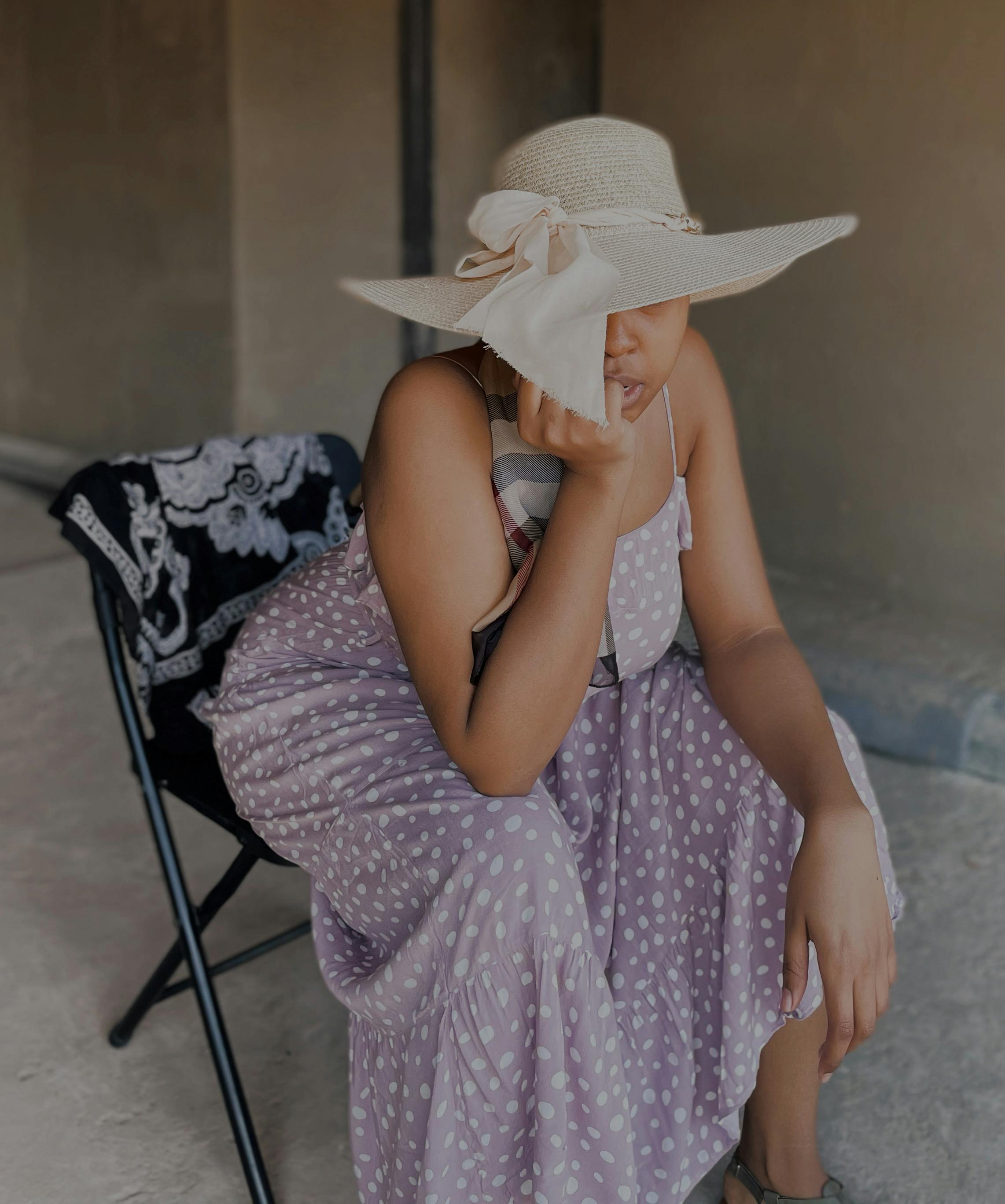 Woman in a polka dot dress and sun hat sitting thoughtfully on a chair indoors.