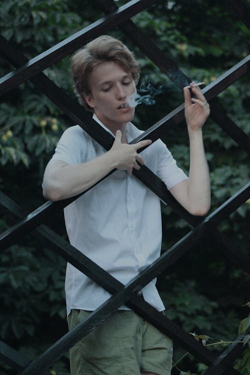 Free A Man in White Shirt Smoking Cigarette Near the Metal Fence Stock Photo