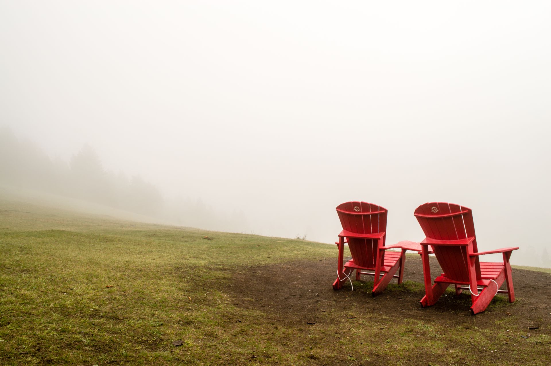 Two red Adirondack chairs on a grassy hill in a foggy landscape, offering a serene outdoor view.