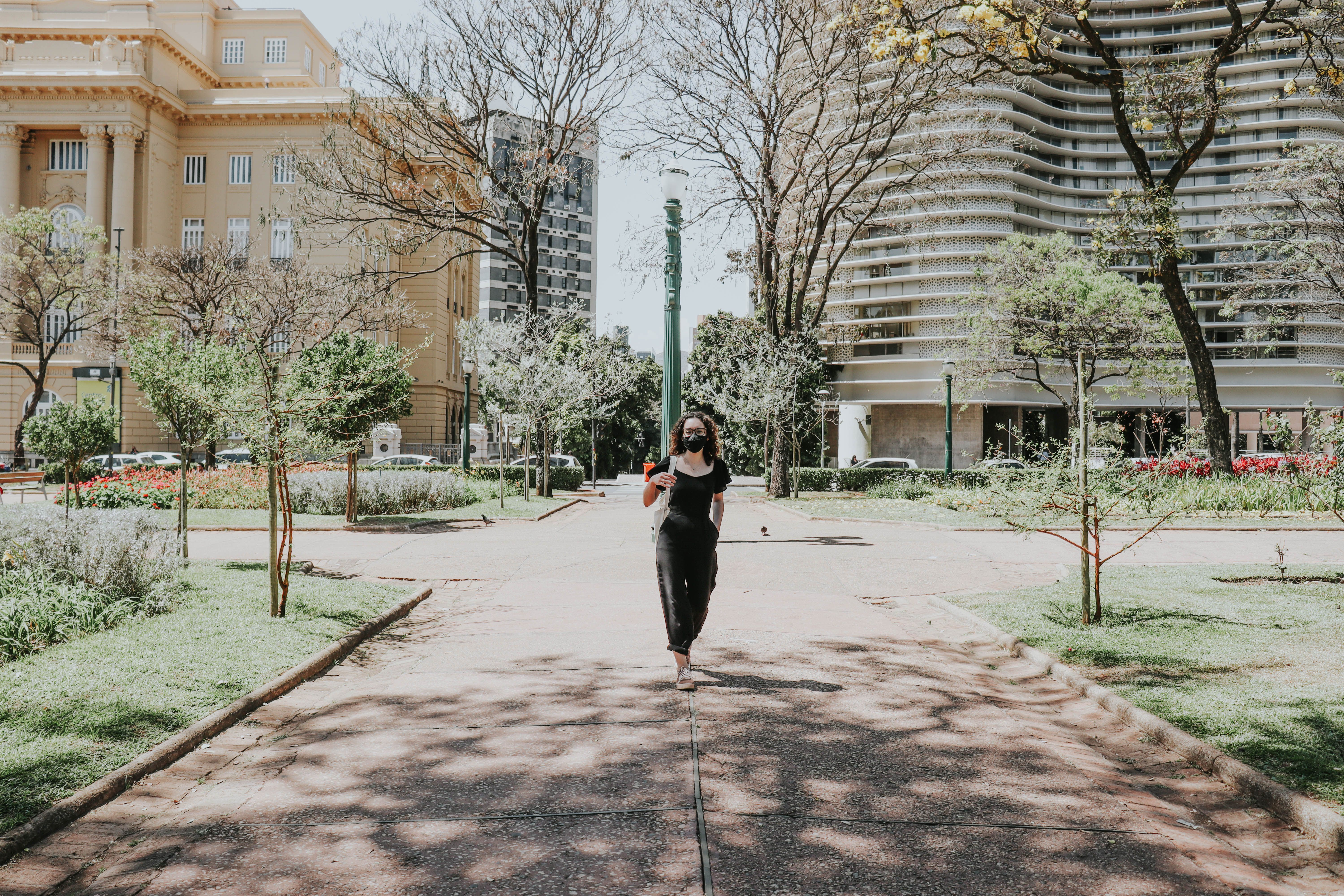A woman in a face mask walks through a sunlit urban park, surrounded by architecture.
