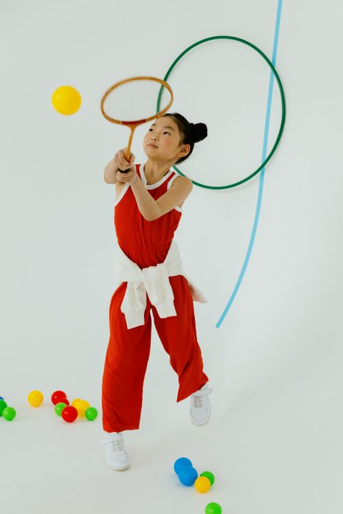 Free A young Asian girl in a red outfit joyfully plays with a racket indoors among colorful balls. Stock Photo
