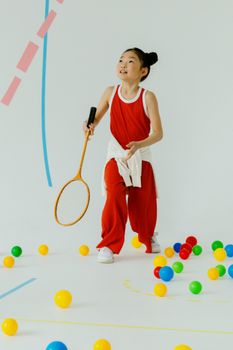 Energetic girl in a red outfit playing badminton indoors surrounded by colorful balls.