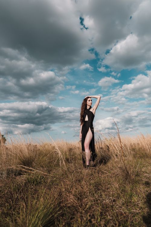 Free Fashionable woman stands in a black dress in a scenic countryside field under a cloudy sky. Stock Photo