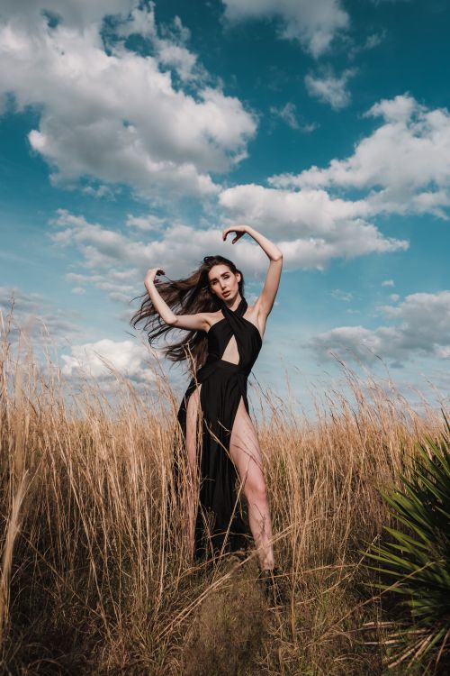 Free Stylish woman posing in black dress, with wind blowing, in Orlando field under a blue, cloudy sky. Stock Photo