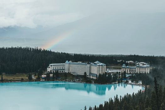 A tranquil lakeside resort surrounded by forest and a rainbow in an overcast sky.