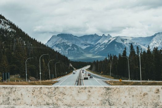 Long road flanked by trees leads to majestic snowcapped mountains under cloudy skies.