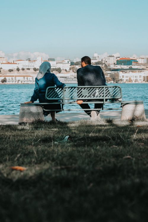 Free Two people sitting on a bench by the waterside, enjoying a cityscape view. Stock Photo