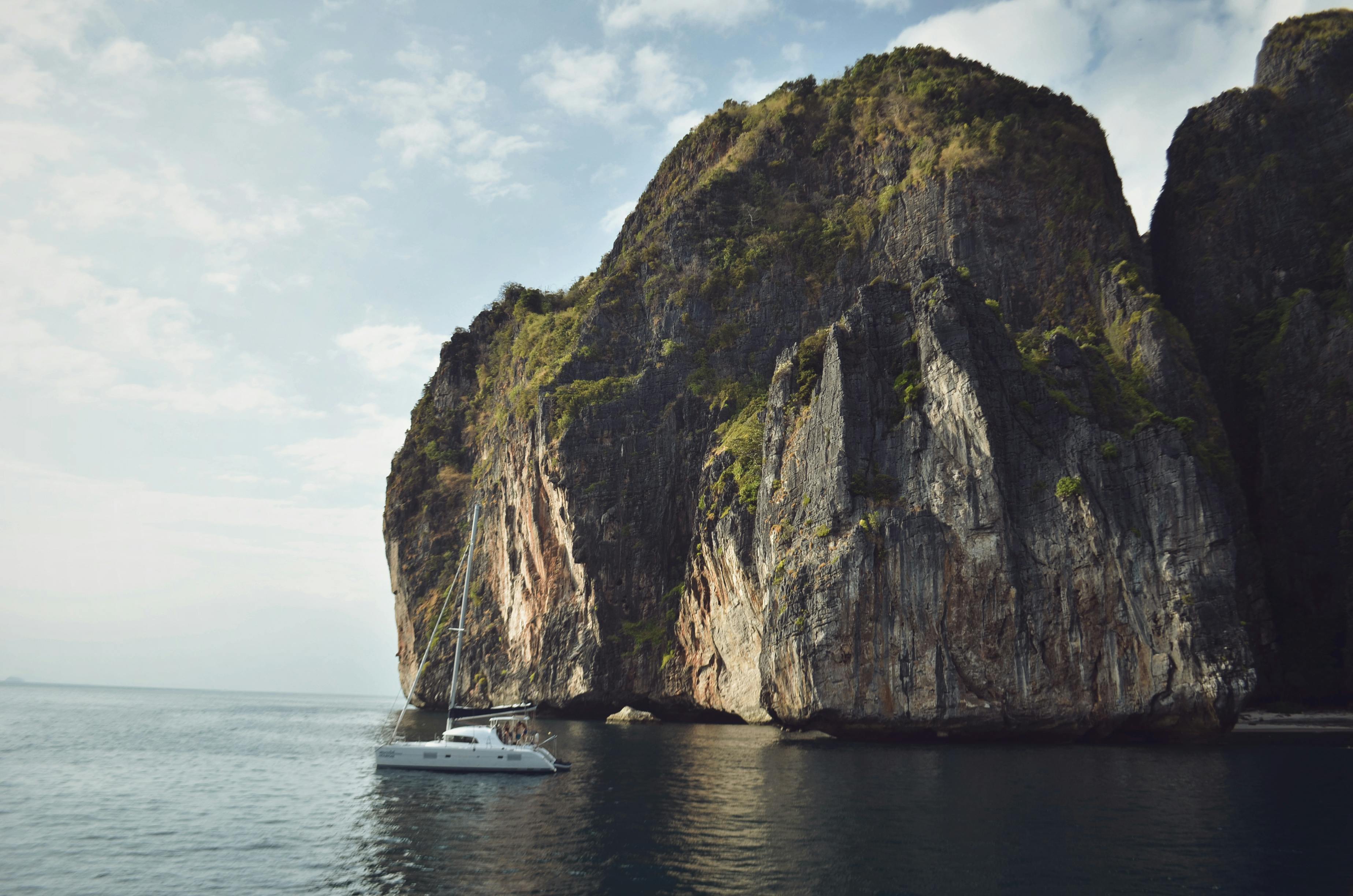 A serene sailboat drifts by a towering island cliff under a cloudy sky.