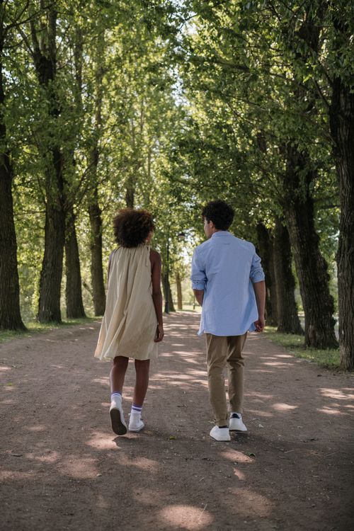 Free A young couple casually walking through a tree-lined park pathway, enjoying a sunny day together. Stock Photo