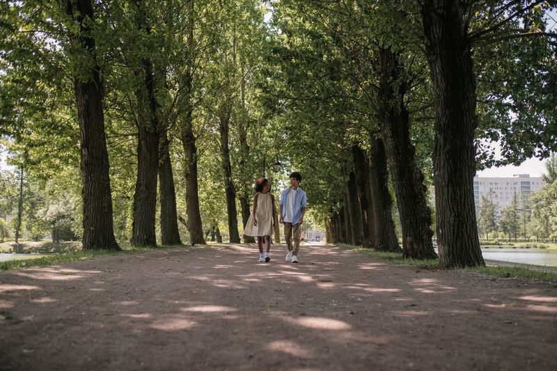 A young couple of teenagers walking under trees on a sunny day in a park, embracing nature and leisure.