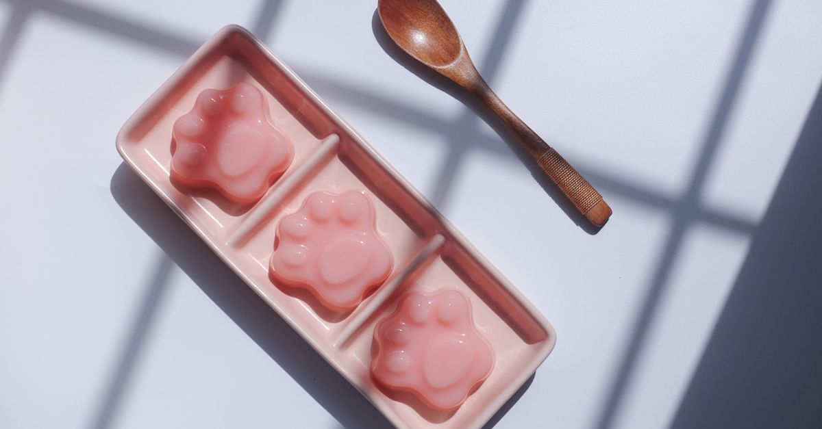 Três gélatinas fofas em forma de patinha em uma bandeja rosa com uma colher de madeira, perfeitas para amantes de sobremesas.