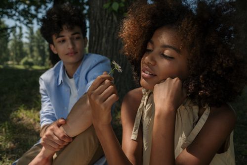 Free Teenagers sitting together in a sunny park, sharing a tender moment with a flower. Stock Photo