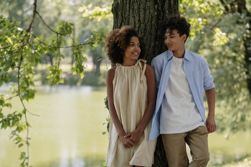 Free A young couple with afro hair enjoying a sunny day by the lake, smiling and leaning against a tree. Stock Photo