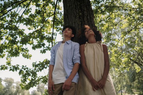 Free Two teenagers leaning against a tree enjoying a sunny day in a park. Captures friendship and relaxation. Stock Photo