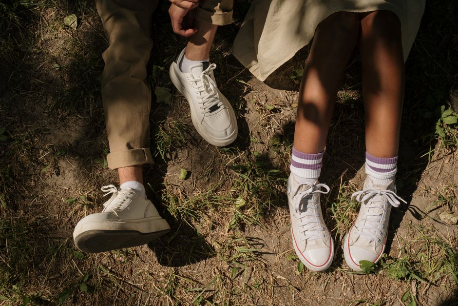 Close-up of two people sitting on grass, focusing on their legs and shoes, in a sunny outdoor setting.
