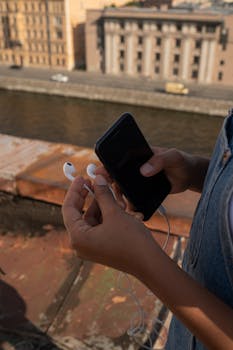 Close-up of hands with smartphone and earbuds on a sunny rooftop near a flowing river.