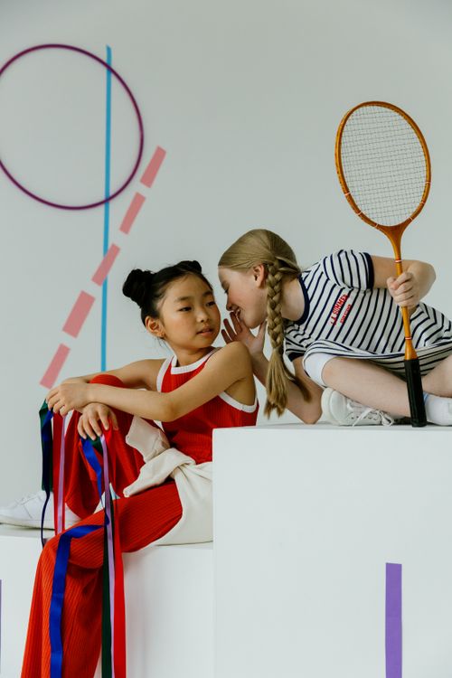 Free Two girls in sportswear whispering, with a badminton racket, set against a playful geometric backdrop. Stock Photo