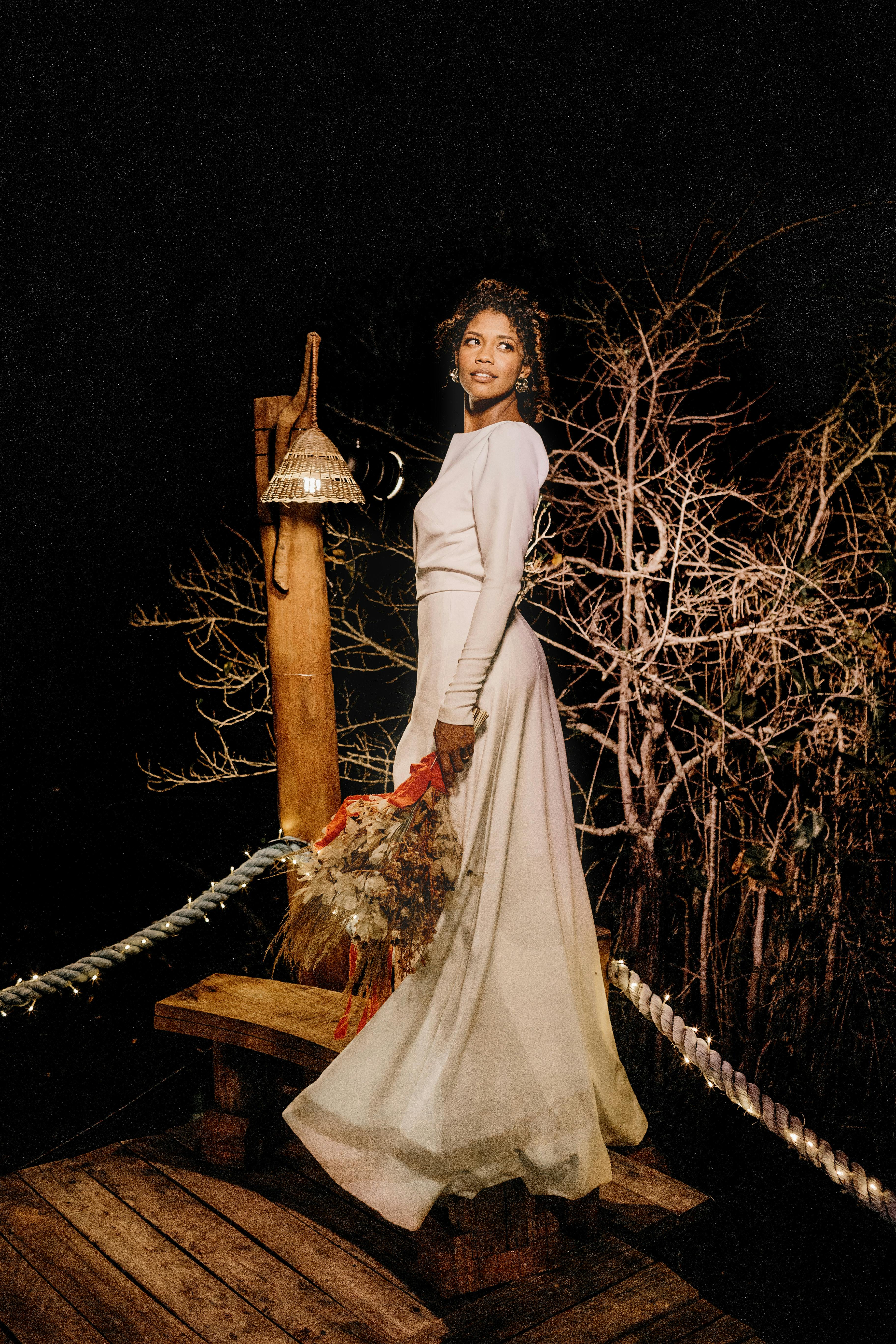 A beautiful bride poses in a long white dress with a bouquet, outdoors at night.