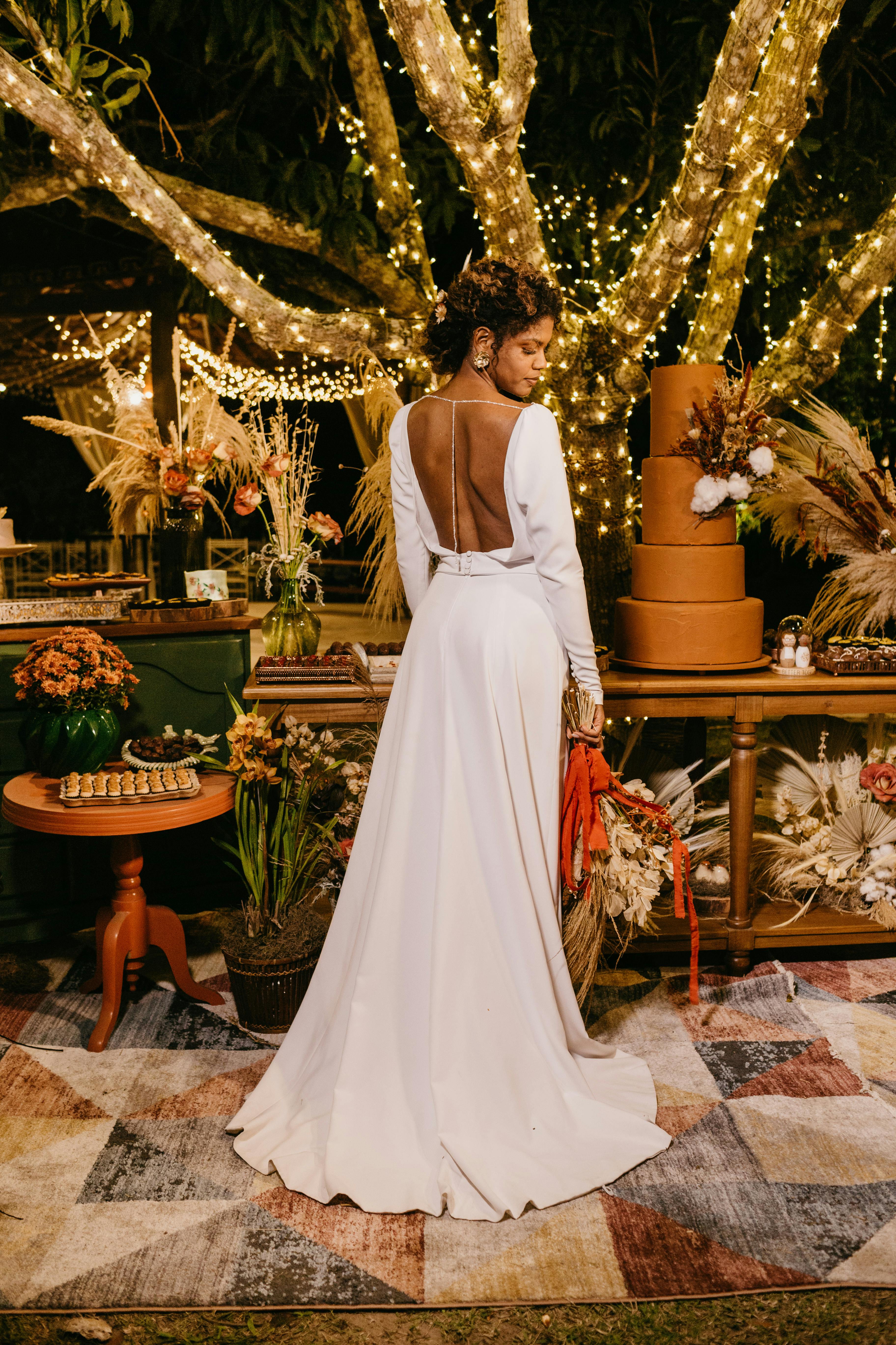 优雅的新娘穿着令人惊叹的白色婚纱，在闪闪发光的树灯下，在户外的花园环境中