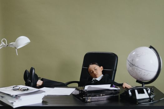 Child in business suit sleeping at desk with office items, indoors, conceptual workplace scene.