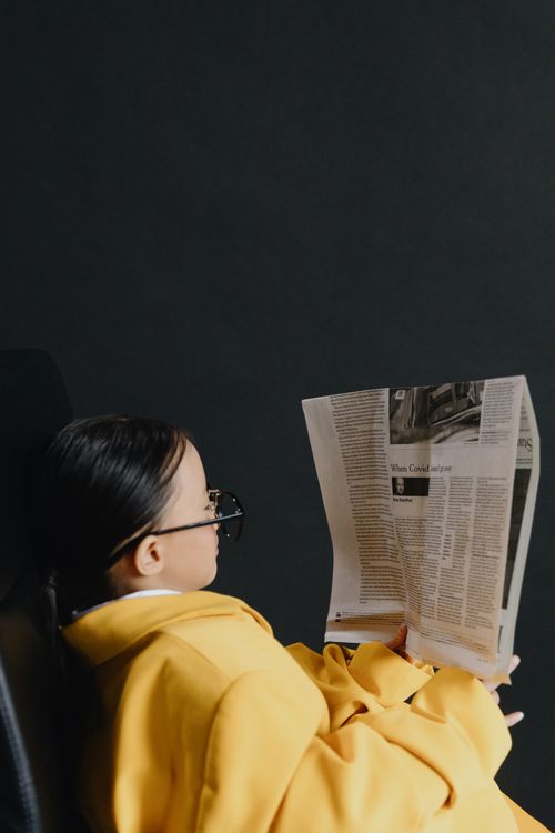 Free A young girl with eyeglasses reads a newspaper indoors, wrapped in a yellow coat. Stock Photo