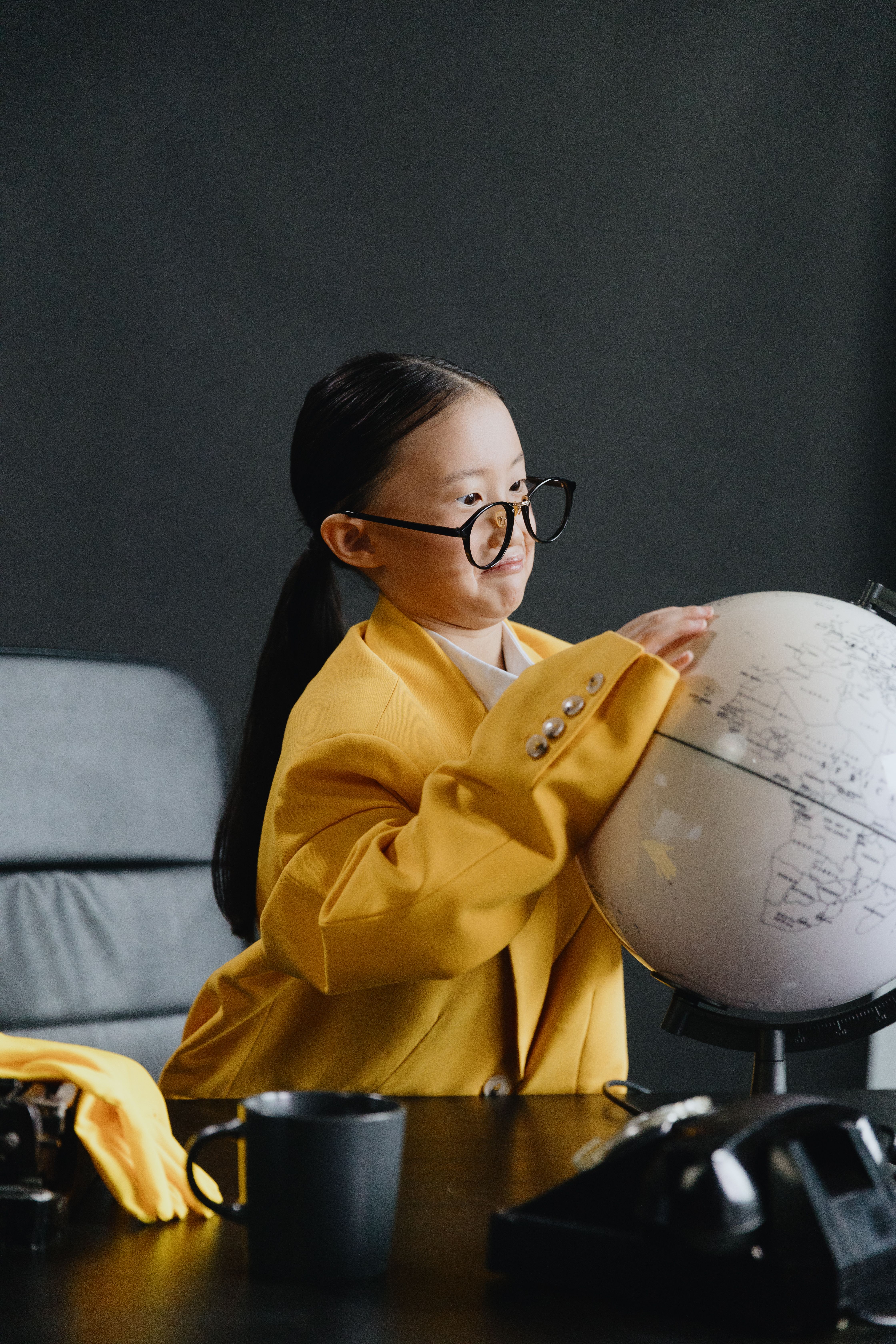 A stylish child wearing a yellow coat and glasses, exploring a globe inside. Fashionable and educational setting.
