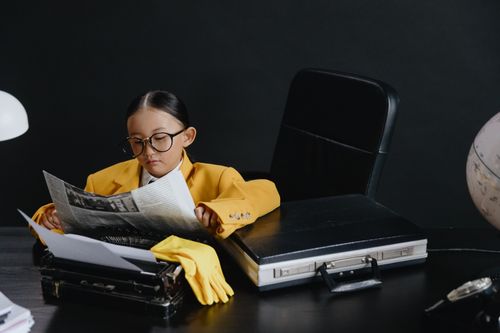 Free A child in eyeglasses and a yellow suit reads a newspaper at a desk, embodying a young journalist. Stock Photo
