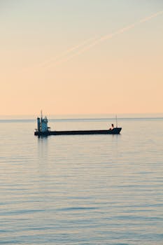 A cargo ship sails across a tranquil sea during a serene sunset, reflecting on the water.