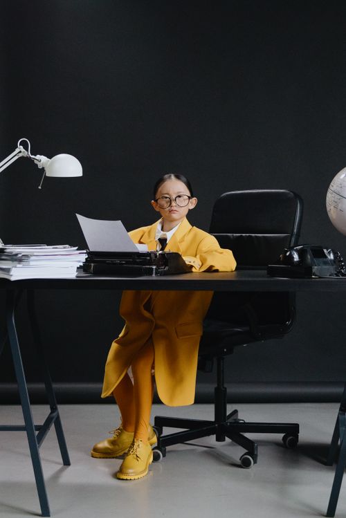 Free Child in yellow coat and eyeglasses sitting at office desk with typewriter and globe. Stock Photo