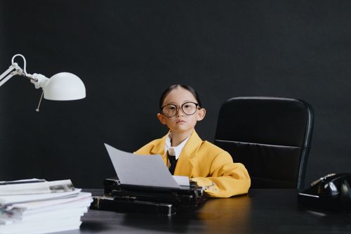 Free Child in yellow blazer with old typewriter, eyeglasses, and lamp at desk suggests business roleplay. Stock Photo