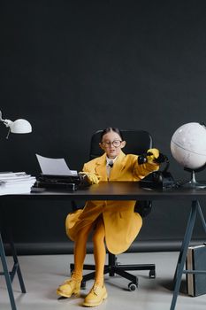 Asian girl in yellow suit with eyeglasses at desk holding a vintage phone with globe nearby.