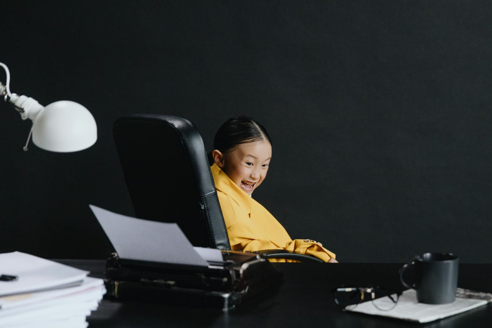 A child joyfully posing in an oversized suit, sitting at an office desk with papers and a lamp.