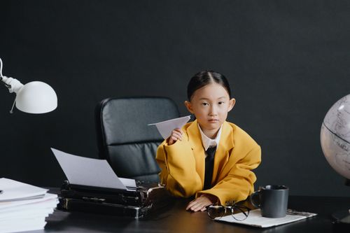 Free A young girl in a yellow jacket sits at an office desk with a paper airplane. Stock Photo