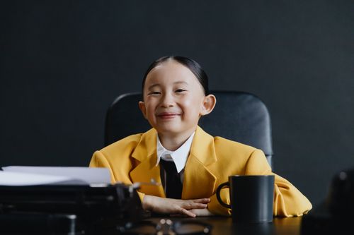 Free A cheerful child wearing a yellow suit smiles while seated at an office desk. Stock Photo