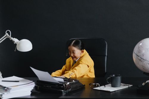 Free A child wearing a yellow suit sits at a desk with a typewriter, looking joyful. Stock Photo