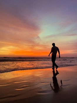 A man walks along the beach, silhouetted against a vibrant sunset, reflecting on the wet sand.